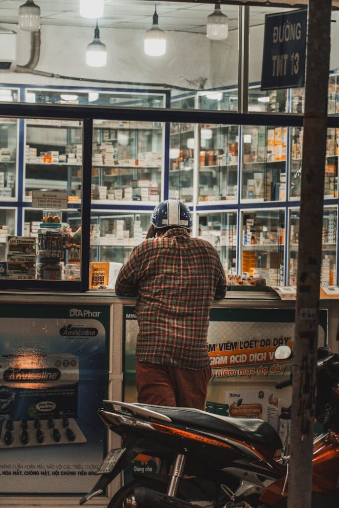 Back view of a man wearing a helmet standing at a pharmacy counter by night in an urban setting.